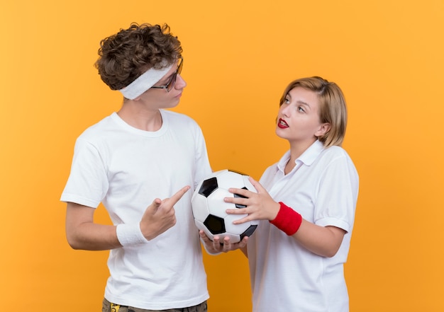 Young sporty couple serious man holding soccer ball looking at his smiling girlfriend standing over orange wall