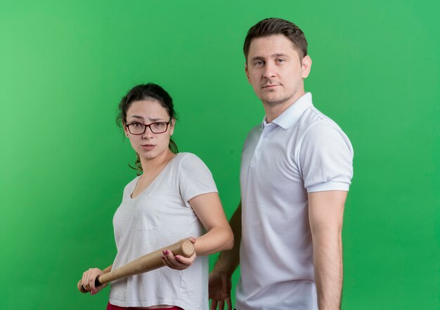 Young sporty couple woman with baseball bat standing next to her boyfriend  with serious face standing over green wall
