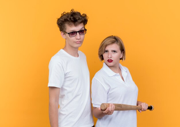 Young sporty couple woman with baseball bat standing next to her boyfriend  with serious face standing over orange wall