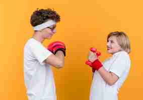 Free photo young sporty couple woman with dumbbells looking at her boyfriend with boxing gloves smiling cheerfully standing over orange wall