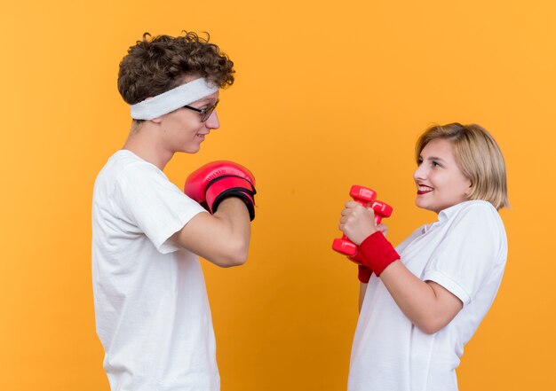 Young sporty couple woman with dumbbells looking at her boyfriend with boxing gloves smiling cheerfully standing over orange wall