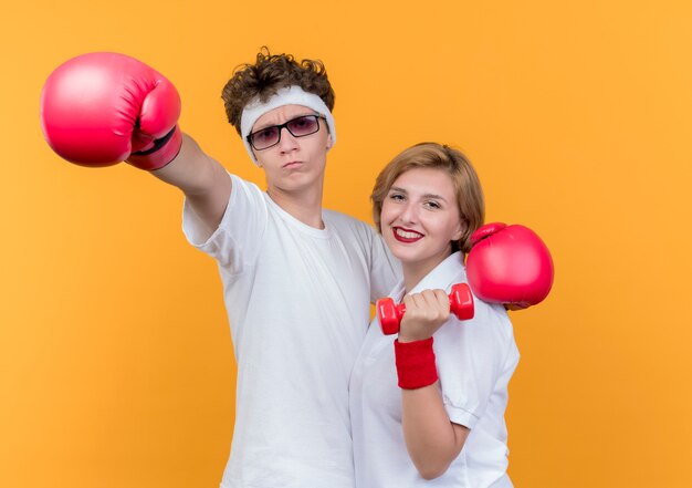 Молодая спортивная пара женщина с гантелями и мужчина с боксерскими перчатками, улыбаясь, стоя над оранжевой стеной