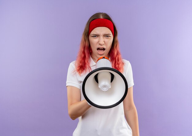 Young sporty girl shouting to megaphone with aggressive expression standing over purple wall