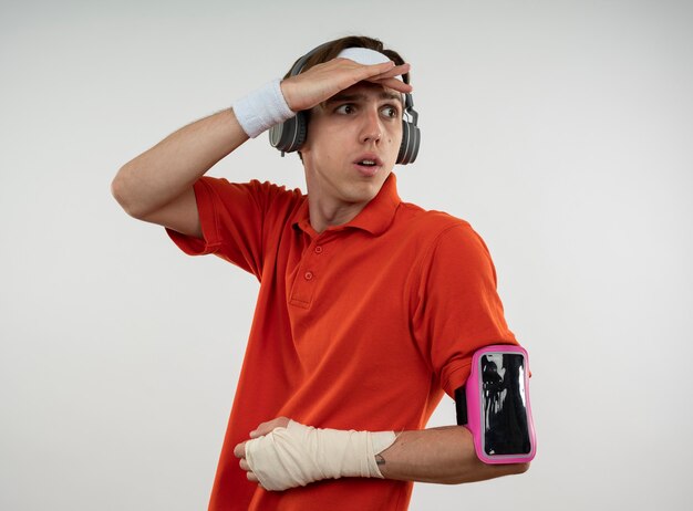 Young sporty guy wearing headband with wristband and headphones with phone arm band looking at distance with hand