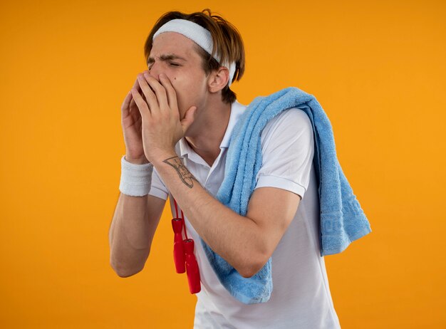 Young sporty guy with closed eyes wearing headband with wristband with jump rope and towel on shoulder calling someone isolated on yellow wall