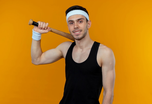 Young sporty man in headband holding basebal bat  smiling confident standing over orange wall