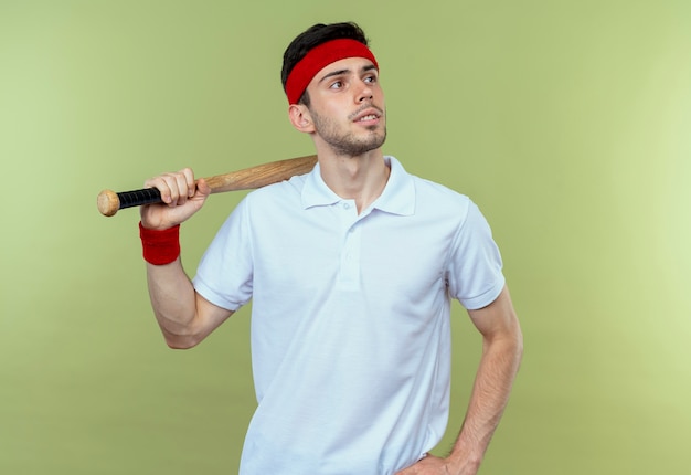 Young sporty man in headband holding baseball bat looking aside with pensive expression standing over green wall