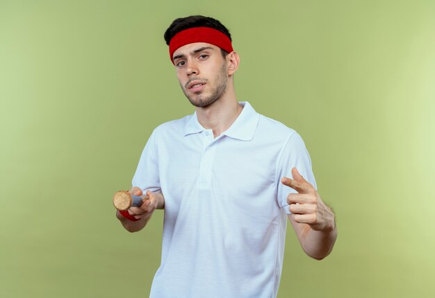 Молодой спортивный мужчина в повязке на голову, держащий бейсбольную биту, указывая пальцем на cemera, выглядит уверенно, стоя над зеленой стеной
