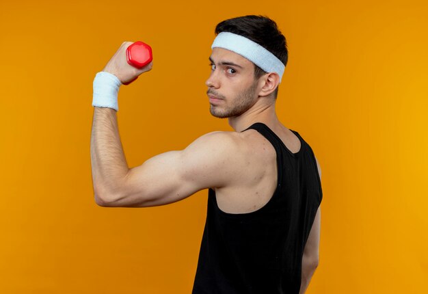 Молодой спортивный мужчина в повязке на голову, держащий гантели, делает упражнения, уверенно выглядит, стоя над оранжевой стеной