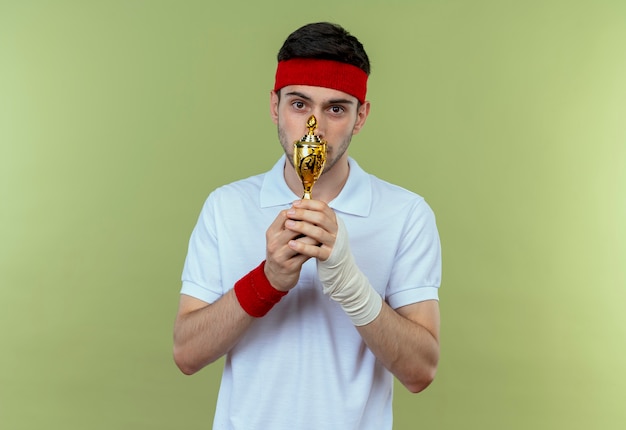 Free photo young sporty man in headband holding his trophy  with shy smile on face standing over green wall