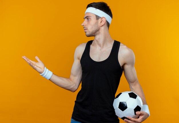 Young sporty man in headband holding soccer ball looking aside with arm out as asking or arguing standing over orange wall