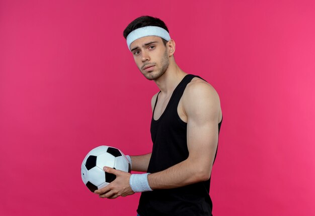 ピンクの背景の上に立っている真剣な表情でカメラを見てサッカーボールを保持しているヘッドバンドの若いスポーティな男