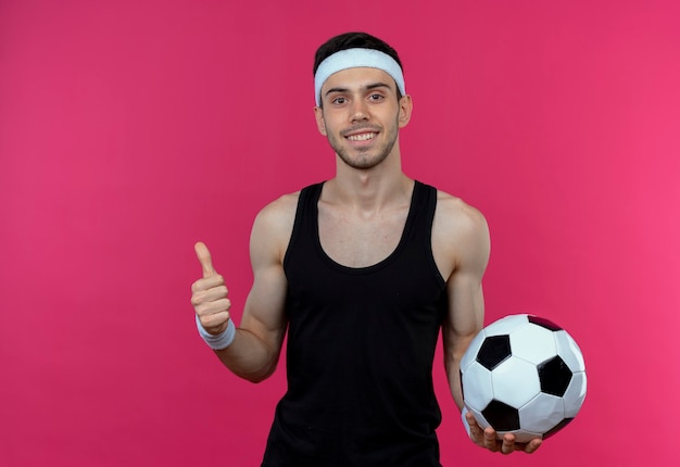 Молодой спортивный мужчина в повязке на голову, держащий футбольный мяч, улыбается, показывает палец вверх, стоя над розовой стеной