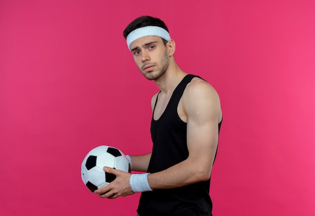 Free photo young sporty man in headband holding soccer ball  with serious expression standing over pink wall