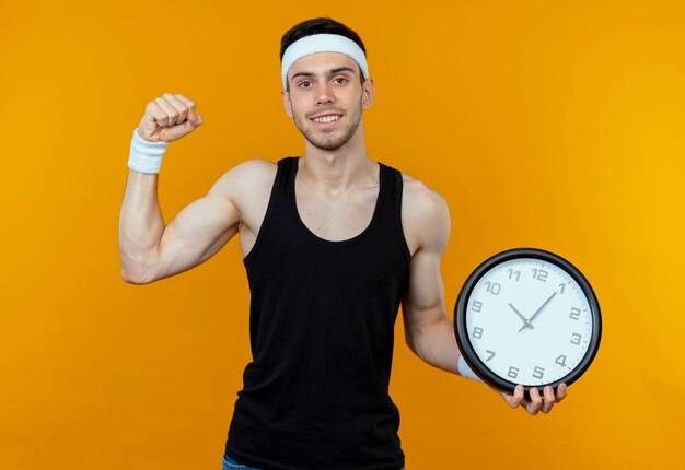 Молодой спортивный мужчина в повязке на голову, держащий настенные часы, сжимая кулак, счастлив и взволнован, стоя на оранжевом фоне