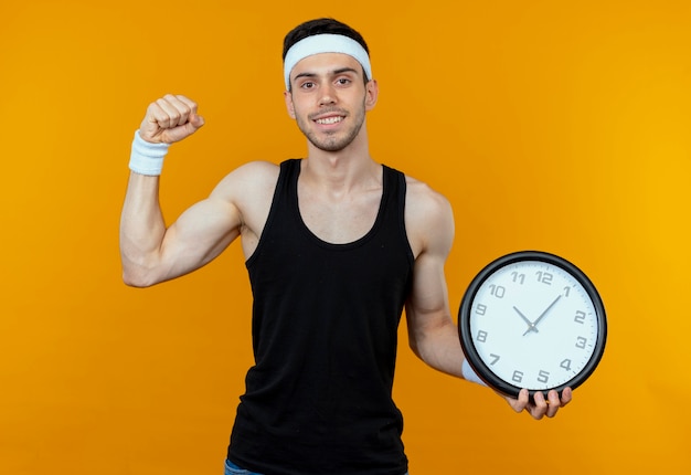 Молодой спортивный мужчина в ободке держит настенные часы, сжимая кулак, счастливый и взволнованный, стоя над оранжевой стеной
