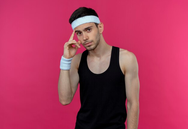Young sporty man in headband looking aside pointing his temple thinking over pink