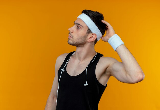 Молодой спортивный мужчина в повязке на голову озадаченно почесывает голову, стоя у оранжевой стены