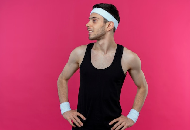 Young sporty man in headband looking aside with arms at hip smiling standing over pink wall