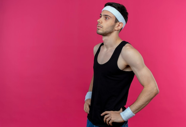 Young sporty man in headband looking aside with serious face standing over pink wall