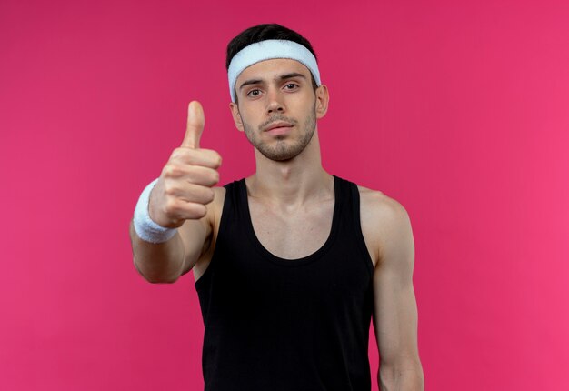 Young sporty man in headband looking at camera showing thumbs up standing over pink background