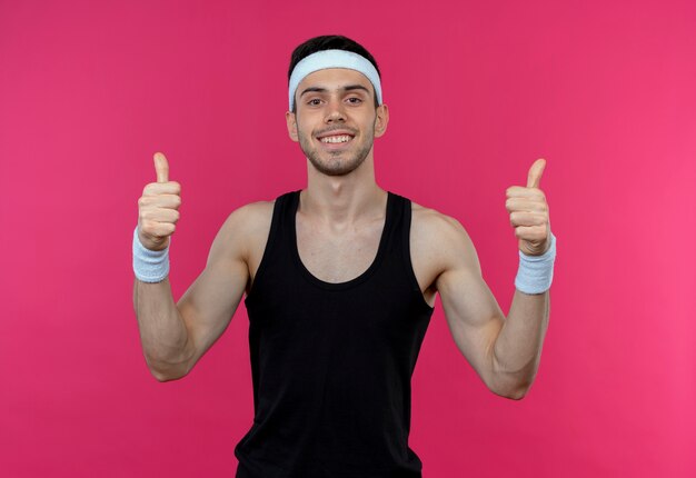 Young sporty man in headband looking at camera smiling happy and positive showing thumbs up standing over pink background