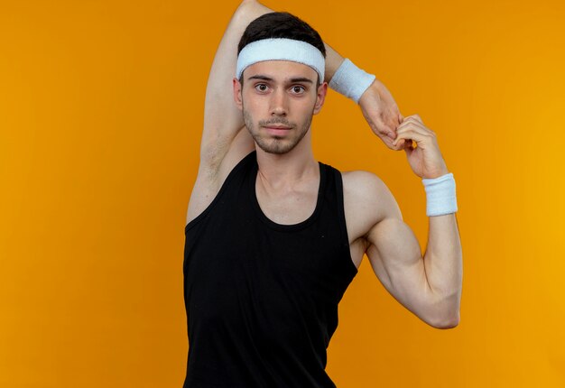 Young sporty man in headband looking at camera stretching his hands standing over orange background