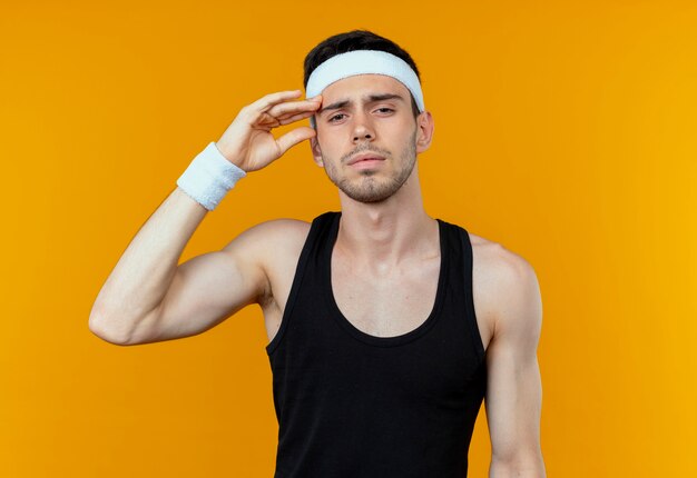 Young sporty man in headband looking at camera touching his head tired and bored standing over orange background