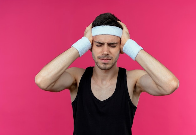 Free photo young sporty man in headband looking unwell holding his head with hands suffering from strong headache standing over pink background