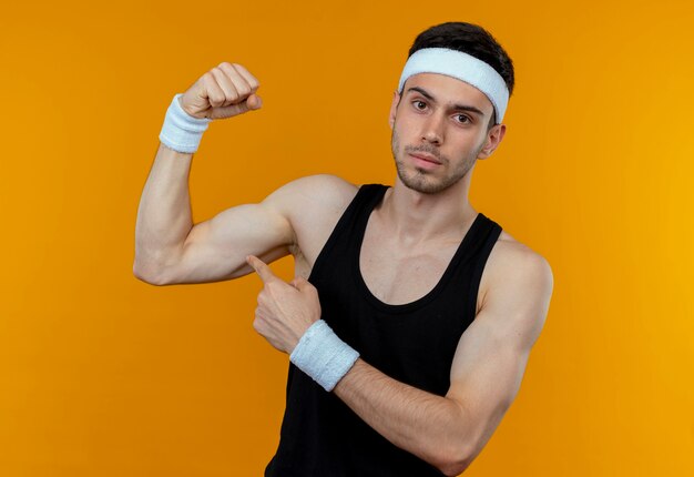 Young sporty man in headband raising fist showing biceps with serious confident expression standing over orange background