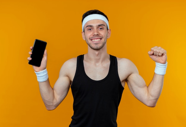 Young sporty man in headband showing smartphone clenching fist happy and excited over orange