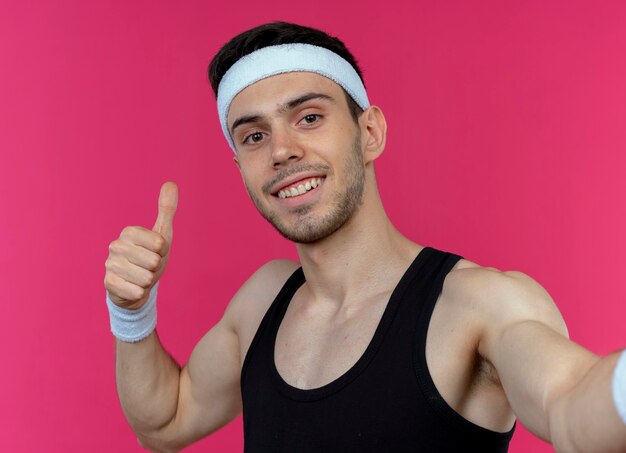 Young sporty man in headband  showing thumbsup smiling standing over pink wall