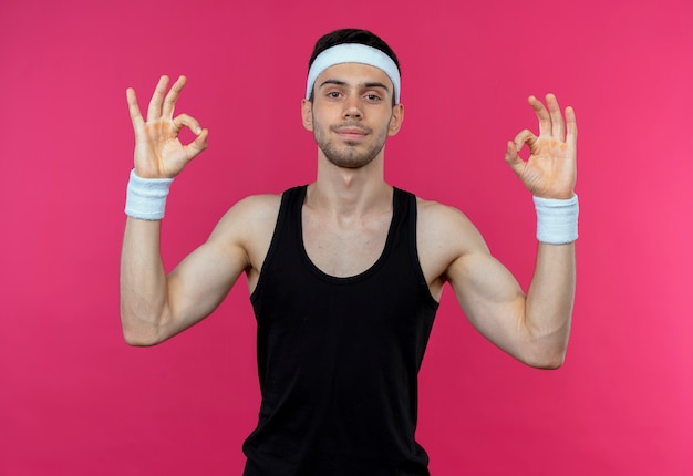 Young sporty man in headband  smiling doing meditation gesture with fingers standing over pink wall