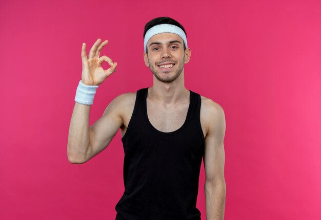 Young sporty man in headband  smiling happy and positive showing ok sign standing over pink wall