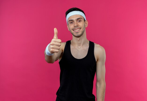 Молодой спортивный мужчина в повязке на голову улыбается счастливым и позитивным, показывает палец вверх над розовым