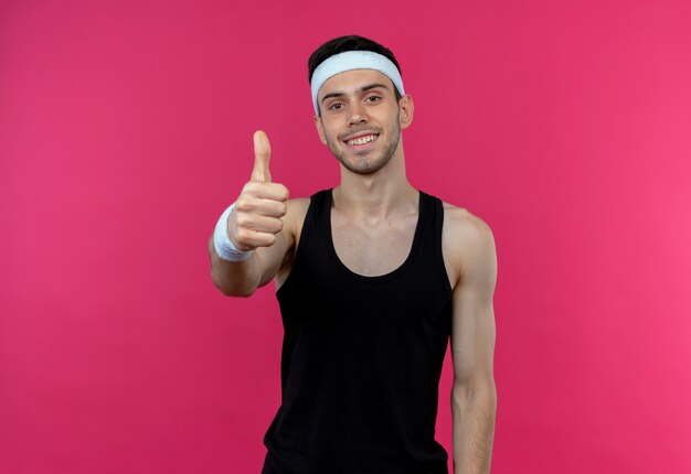 Young sporty man in headband smiling happy and positive showing thumbs up over pink