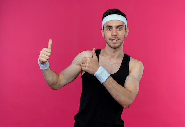Young sporty man in headband  smiling happy and positive showing thumbs up standing over pink wall