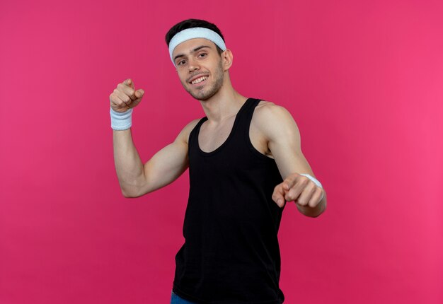 Молодой спортивный мужчина в повязке на голову улыбается, указывая указательными пальцами на камеру над розовым