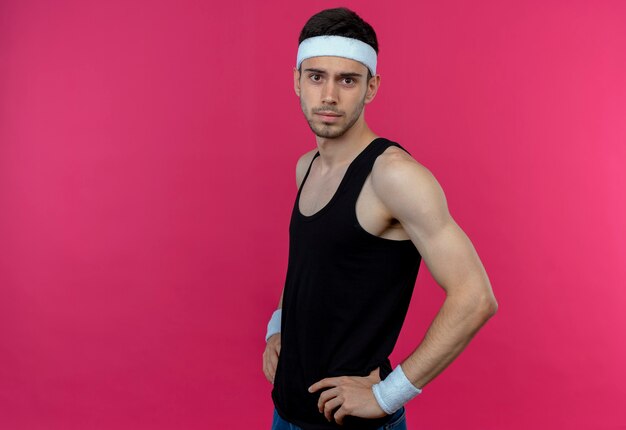 Young sporty man in headband  with confident serious expression with arms at hip standing over pink wall