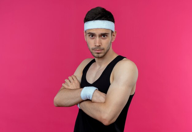 Young sporty man in headband  with confident serious expression with crossed arms on chest standing over pink wall