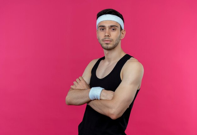 Молодой спортивный мужчина в повязке на голову с уверенным серьезным выражением лица со скрещенными руками на груди, стоящий над розовой стеной