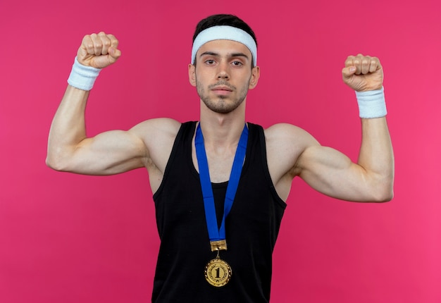 Young sporty man in headband with gold medal around neck looking at camera raising fist with serious expression standing over pink background