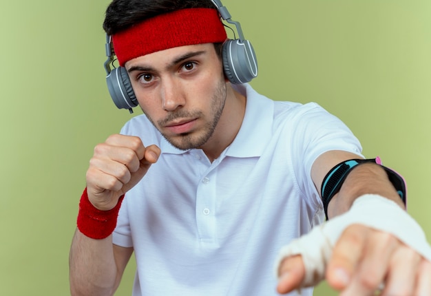 Giovane uomo sportivo in fascia con cuffie e fascia da braccio per smartphone in posa come un combattente con il pugno chiuso sul verde