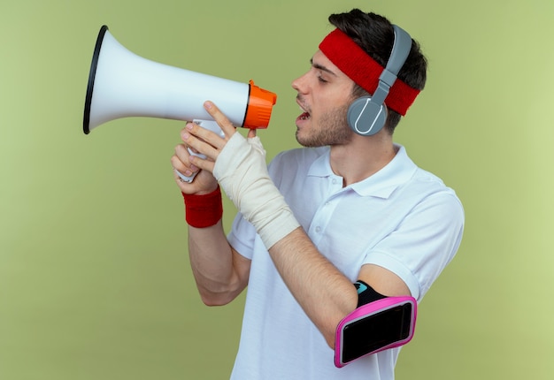 Young sporty man in headband with headphones and smartphone arm band shouting through megaphone standing over green background