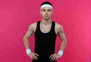 Free photo young sporty man in headband with serious face with arms at hip over pink