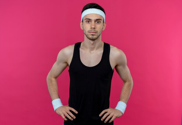 Young sporty man in headband with serious face with arms at hip over pink