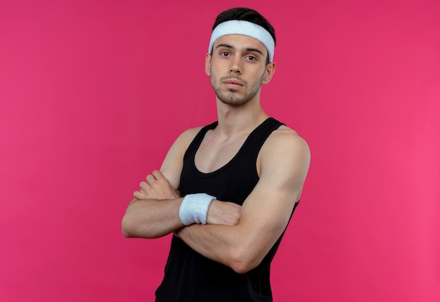 Young sporty man in headband with serious face with crossed arms on chest over pink