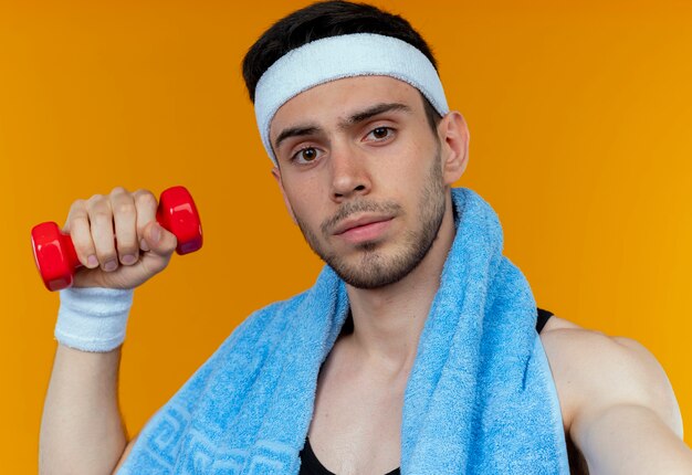 Young sporty man in headband with towel around neck holding dumbbel in raised hand with serious face over orange