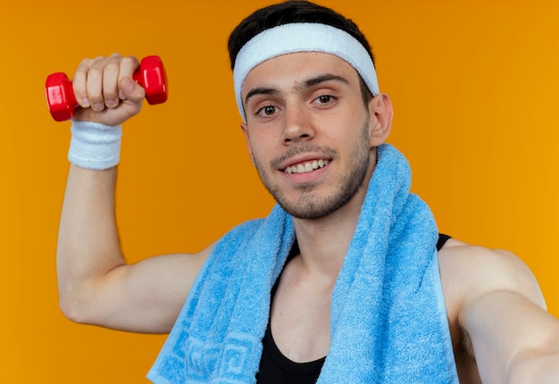 Young sporty man in headband with towel around neck holding dumbbell looking at camera with smile on face standing over orange background