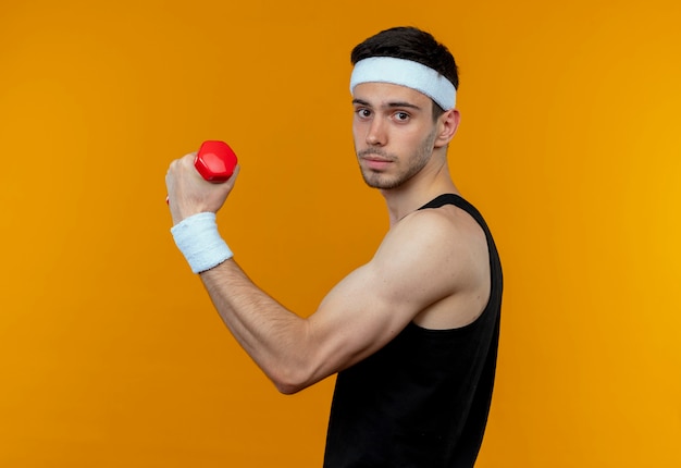 Young sporty man in headband working out with dumbbell looking at camera with serious face standing over orange background